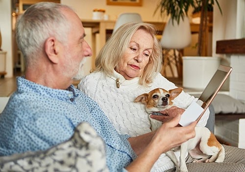 An elderly couple on the couch with their dog, researching social security benefits on their tablet.