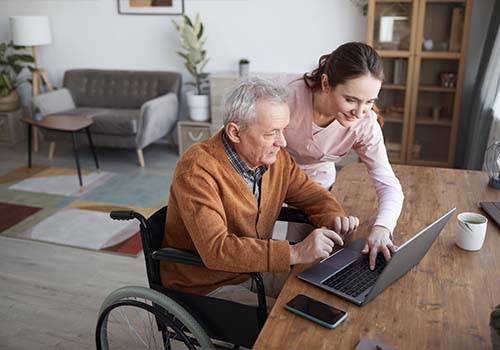 Portrait Of Senior Man In Wheelchair Using Laptop With Nurse Helping Him