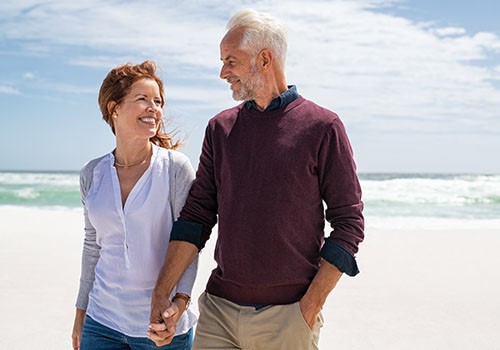 A retired couple walking on the beach smiling at each other.