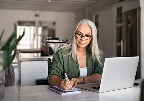 Senior Woman Taking Notes While Using Laptop At Home