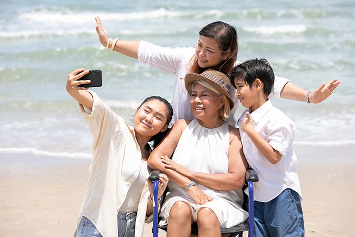 A disabled woman in a wheelchair, surrounded by her family on a sunny beach taking a selfie.