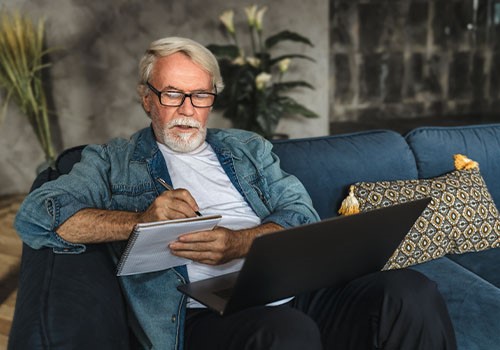 An elderly man sitting on a couch with his laptop while calculating his social security benefits.