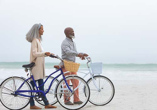 Side View Of Retired Couple Walking With Bicycles On Beach