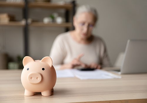 A piggy bank in the foreground and an elderly woman using a calculator and computer in the background.