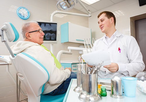 An elderly man sits in the dentist chair as the dentist talks to him.