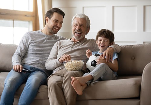 An elderly man, a young man, and a child are sitting on a couch, laughing and eating popcorn.