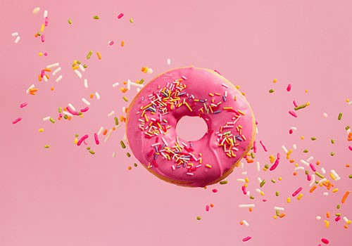 A pink glazed donut flying through the air with sprinkles flying off it.