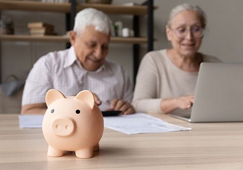 An elderly couple sitting at a desk with a laptop and documents with a piggy bank in the foreground.